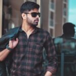 Trendy man with beard wearing sunglasses and plaid shirt, holding a leather jacket outdoors.